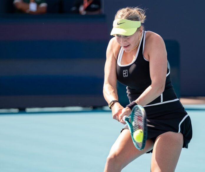 Amanda Anisimova hits a two-handed backhand during a match at the 2026 Miami Open, wearing a black dress and lime-yellow visor, eyes fixed on the ball.