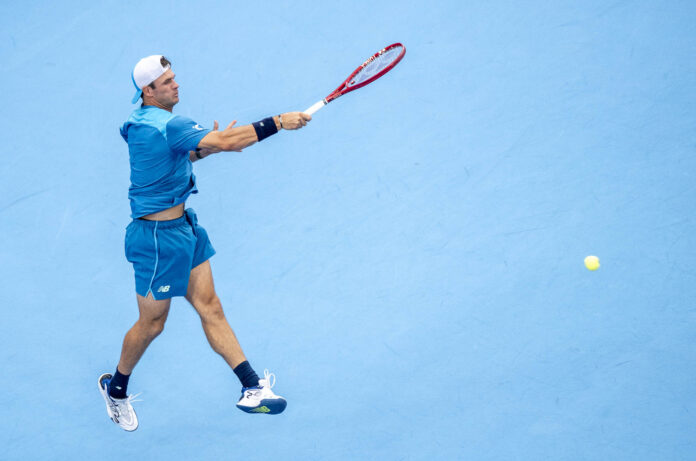Tommy Paul stretches to hit a return on a blue hard court, photographed from high above, with the ball in flight to his right.