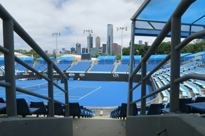 An empty blue hard tennis court surrounded by empty stadium seating, with a city skyline behind